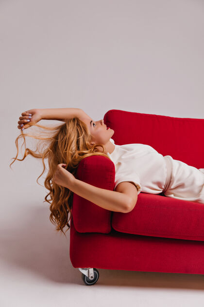 迷人梦幻般的女人躺在红沙发上仰望沉思的金发女孩抚摸着她的头发女人寒冷沙发