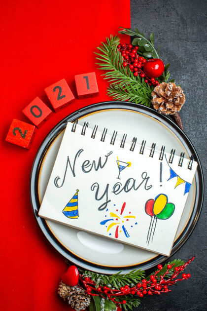 抽签新年背景的垂直视图 笔记本上有新年图纸 餐盘上有装饰配件 杉木树枝和数字放在黑色桌子上的红色餐巾上笔记本圣诞节餐巾