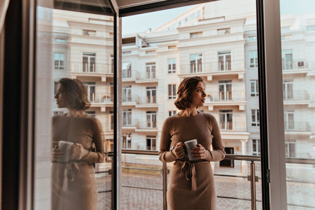 阴影身着棕色连衣裙的沉思的女人在喝卡布奇诺阳台边站着一杯咖啡的漂亮女孩的画像放松灯光毛衣