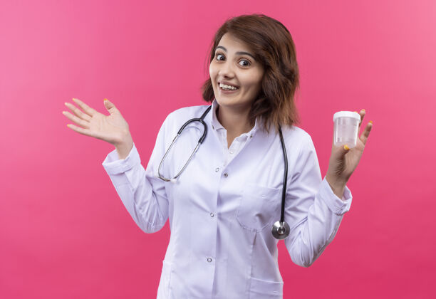 测试穿着白大褂的年轻女医生拿着听诊器 拿着测试罐 面带困惑 微笑着把胳膊伸到一边站着看听诊器