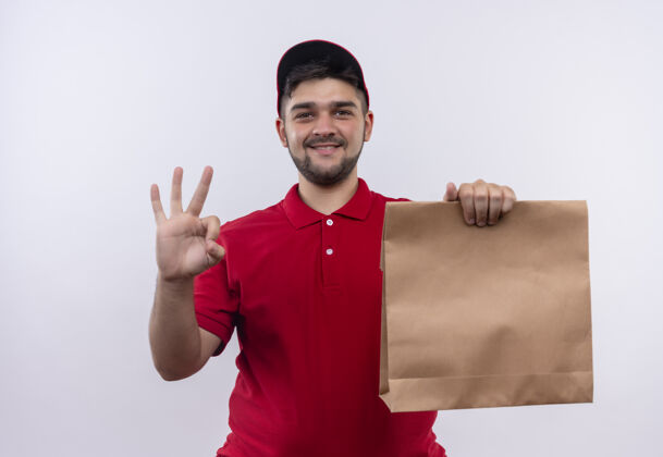 纸年轻的送货员穿着红色制服 戴着帽子 手里拿着纸包 微笑着友好地展示着ok标志帽子男孩红色