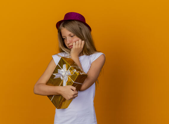 盒子戴着节日礼帽的漂亮小女孩拿着礼盒边看边用沉思的表情思考 生日聚会的概念站在橙色的背景上思考帽子小