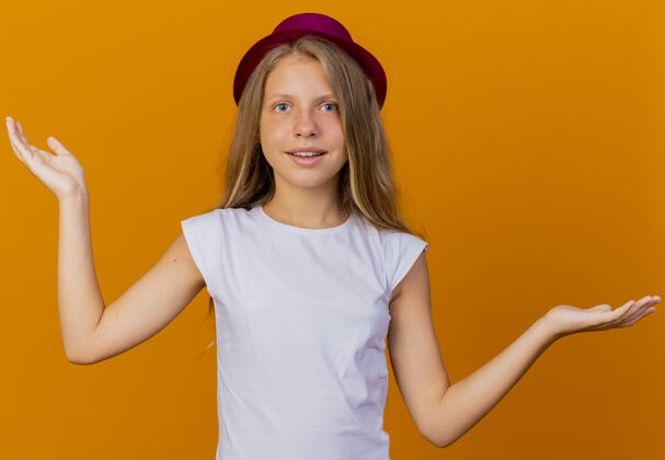 胳膊戴着节日礼帽的漂亮小女孩看着相机微笑着向两边张开双臂 橙色背景下站着生日派对的概念壁板小节日