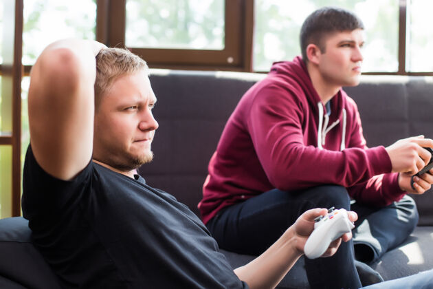 啤酒两个快乐的年轻人坐在沙发上玩电子游戏微笑成功操纵杆