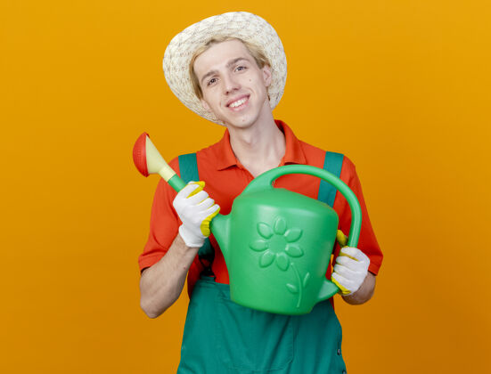 浇水快乐的年轻园丁 穿着连体衣 戴着帽子 拿着喷壶 站在橙色的背景上 看着摄像机 开心地笑着橙色年轻相机