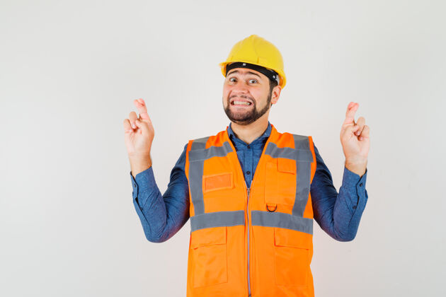 衬衫年轻的建筑工人穿着衬衫 背心 头盔保持手指交叉 看起来很高兴 前视图工作工程师男性