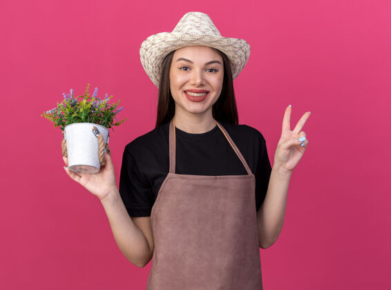 园艺微笑着美丽的白人女园丁戴着园艺帽拿着花盆和胜利的手势标志隔离在粉红色的墙上复制空间女帽子花园