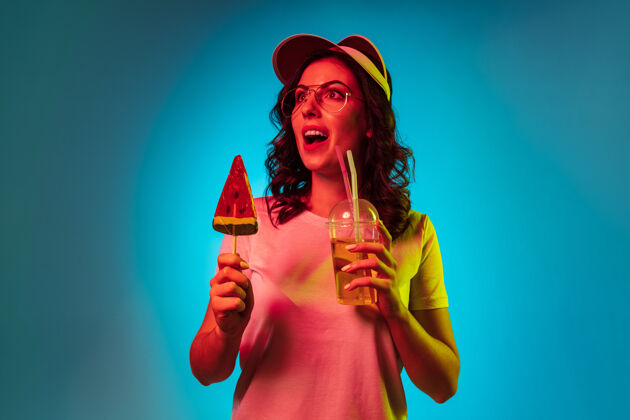 霓虹灯快乐的年轻女子戴着帽子拿着糖果和饮料在时髦的蓝色霓虹灯工作室女性站派对