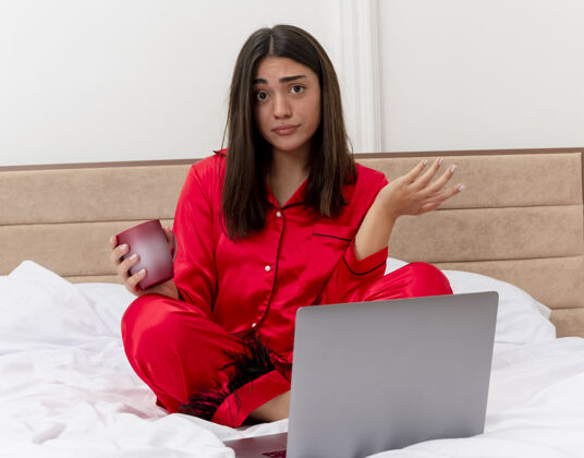 笔记本电脑穿着红色睡衣的年轻美女坐在床上 拿着笔记本电脑和一杯咖啡看着摄像机 困惑地将手臂伸向卧室内部灯光背景下的一侧看手臂年轻