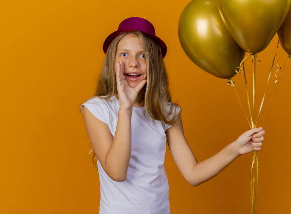 嘴巴戴着节日礼帽的漂亮小女孩 手放在嘴边 手里拿着一堆巴龙 站在橙色的背景下 高喊着生日派对的概念束巴龙生日