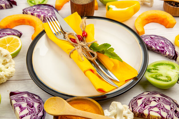 盘子底视图把叉子和刀子绑在黄色餐巾上放在白色盘子上切蔬菜红色卷心菜南瓜花椰菜黄色甜椒放在白色木桌上绑好卷心菜餐巾
