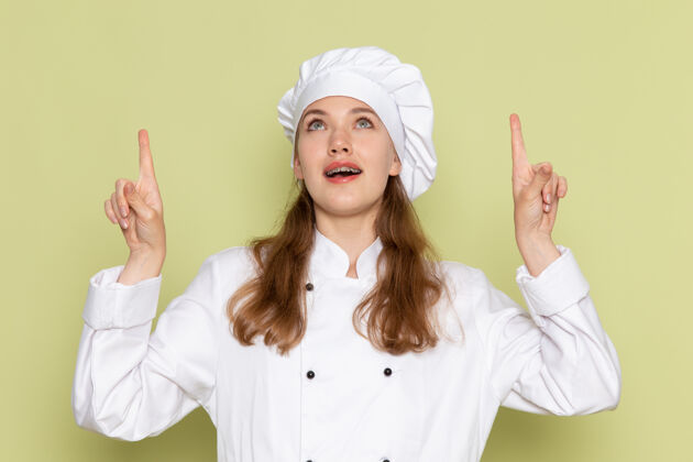 人身着白色厨师服的女厨师在绿色墙壁上抬起手指摆姿势的正视图烹饪姿势西装