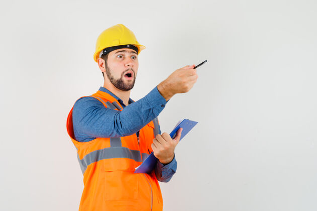 建筑工人年轻的建筑工人穿着衬衫 背心 头盔 一边拿着剪贴板 一边给出指示工人建筑剪贴板