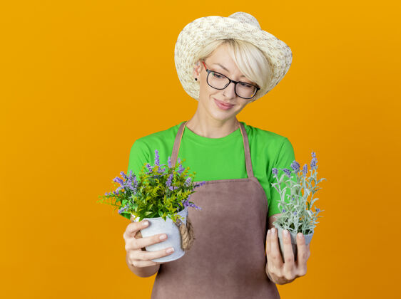 女人一位年轻的园丁 围裙上留着短发 戴着帽子 手里拿着盆栽植物 站在橙色的背景上 面带微笑地看着它们橙色年轻帽子