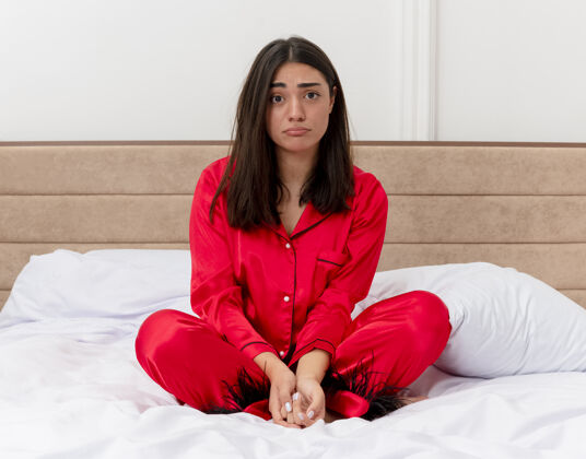 年轻穿着红色睡衣的年轻漂亮女人坐在床上看着相机 在灯光背景下的卧室内部表情悲伤表情相机床
