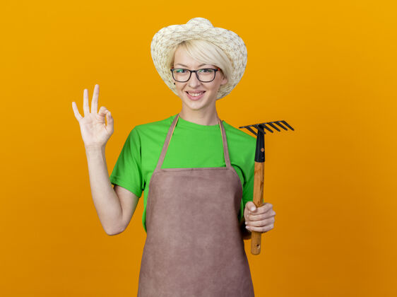 标志一位年轻的园丁 围裙上留着短发 戴着帽子 手里拿着小耙子 微笑着看着摄像机 橙色背景上立着“ok”的牌子帽子看Ok
