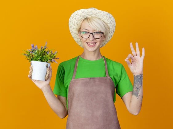 看一位年轻的园丁 围裙上留着短发 戴着帽子 手里拿着盆栽植物 面带微笑地看着摄像机 在橙色的背景上站着一个“ok”的标志短裤花园脸