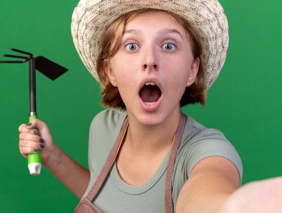 斯拉夫震惊年轻的斯拉夫女园丁戴着园艺帽拿着锄头耙子 在绿色的墙上与复制空间隔离自拍自拍耙采取