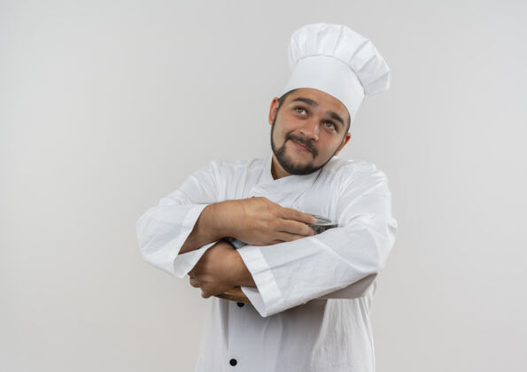 看着高兴的年轻男厨师穿着厨师制服拿着锅碗瓢盆 孤立地抬头望着白色的空间年轻抱着厨师