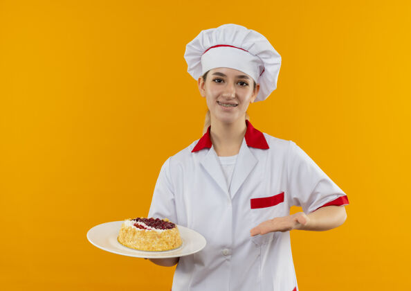 厨师微笑着的年轻漂亮厨师 身着厨师制服 戴着牙套 手拿着并指着孤立在橙色空间的蛋糕盘拿着牙科年轻