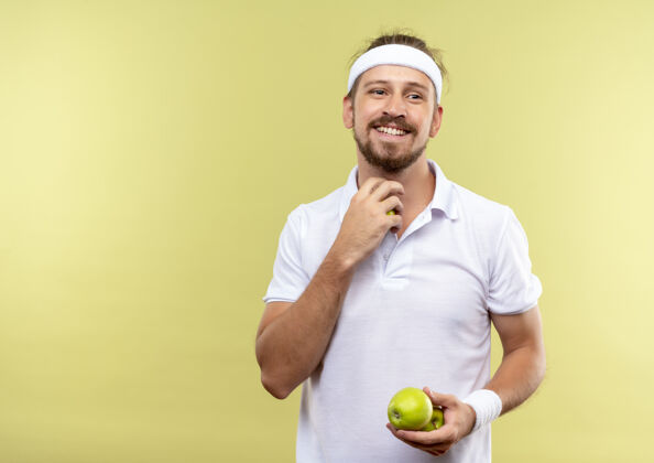 空间面带微笑的年轻帅气的运动型男人戴着头带和手镯拿着苹果看着隔离在绿色空间的一边抱着帅气运动