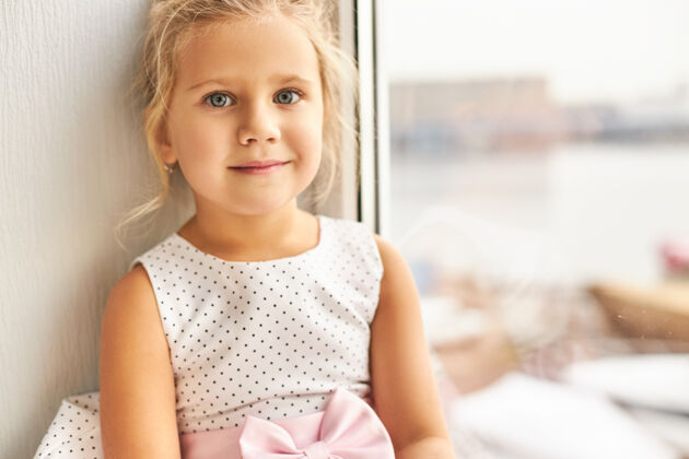 微笑童年和天真的概念迷人可爱的小女孩的肖像 聚在一起的金发和美丽的大眼睛坐在窗前有愉快的表情和微笑玻璃窗台童年