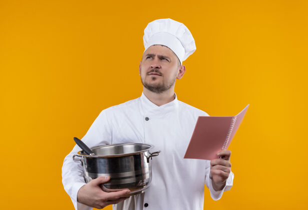 橙色体贴的年轻帅哥厨师穿着厨师制服拿着锅炉和便笺簿仰望孤立的橙色空间年轻厨师垫着