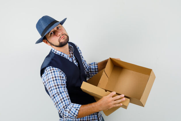 胡茬年轻人展示着空纸箱 穿着衬衫 背心 戴着帽子 看上去很悲伤成功成功商人