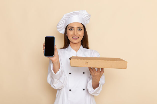 套装身穿白色厨师服的年轻女厨师的正视图 在浅白的墙上拿着电话和食物盒烹饪电话工作