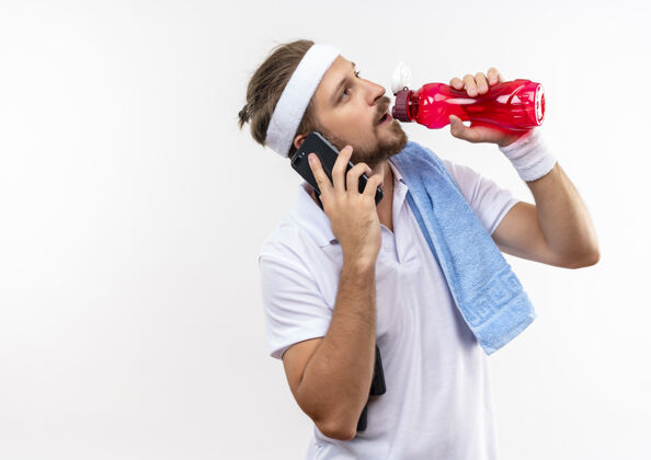 年轻年轻帅气的运动型男人戴着头带和腕带打电话 肩上拿着水瓶 跳绳和毛巾隔离在空白处肩膀电话瓶子
