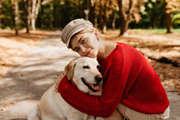 动物可爱的年轻女子戴着漂亮的浅色帽子 穿着红色毛衣 和拉布拉多一起坐在秋天的公园里漂亮的金发女郎和她的狗坐在落叶中宠物金发拉布拉多