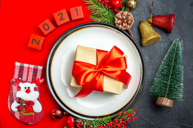 餐盘新年背景俯视图 餐盘上有红丝带装饰配件杉木树枝和数字圣诞袜放在黑色桌子上圣诞树旁的红色餐巾上上衣树枝西红柿