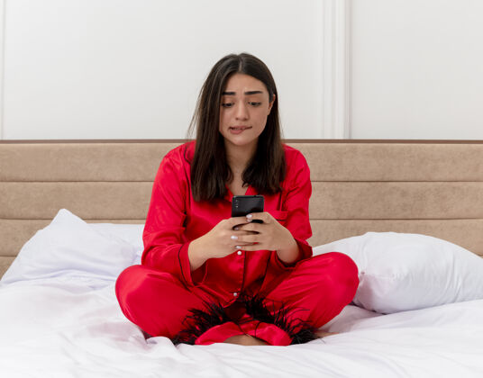 红色穿着红色睡衣的年轻美女坐在床上用智能手机看着卧室里灯光背景下的困惑和紧张坐着卧室紧张