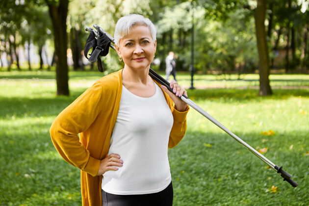 徒步旅行退休后的运动型短发女士对健康积极的生活方式表示赞同 肩上扛着北欧行走的拐杖 准备好好散步 锻炼身体和心血管系统退休棍棒放松