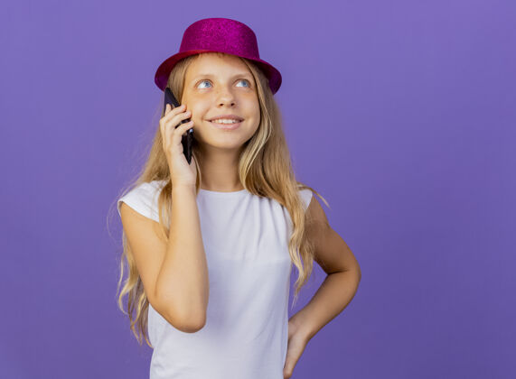 漂亮戴着节日礼帽的漂亮小女孩微笑着用手机交谈 紫色背景下站着生日派对的概念说话女孩帽子