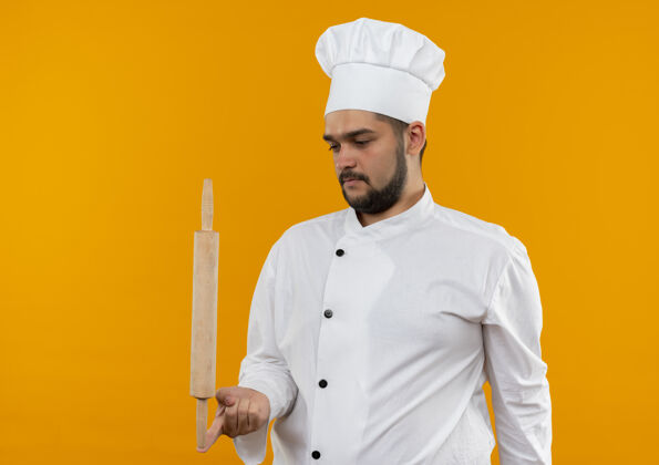 手指年轻的男厨师穿着厨师制服 手指上拿着擀面杖 孤立地看着橙色空间里的擀面杖橙色厨师年轻