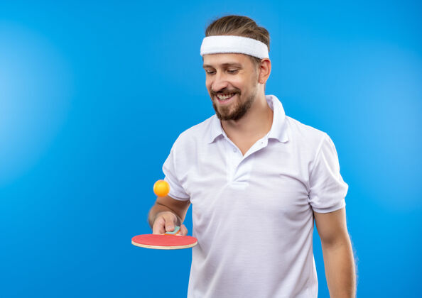 微笑戴着头带和腕带的年轻帅气的运动型男子微笑着用乒乓球拍扔球 孤立地看着它在蓝色的空间里空间球看