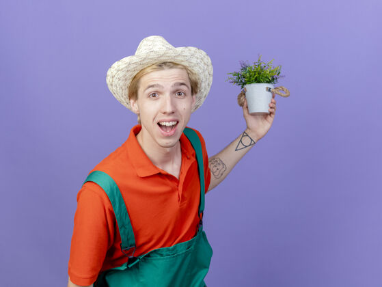脸年轻的园丁 穿着连身衣 戴着帽子 手里拿着盆栽植物 站在紫色的背景上 面带微笑地看着镜头植物连身衣锅