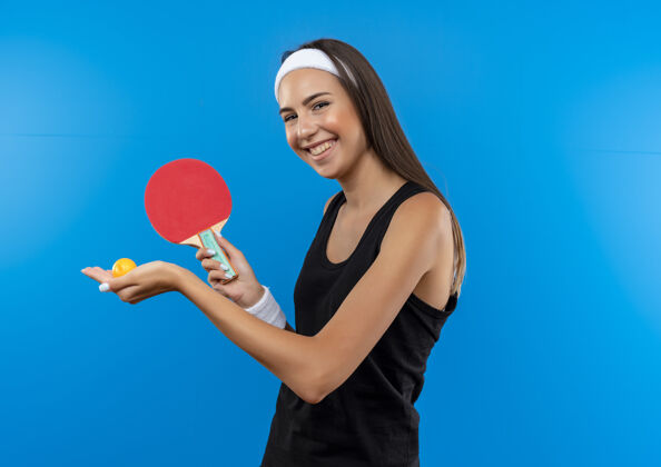 蓝色微笑着的年轻漂亮的运动女孩戴着头带和腕带拿着乒乓球拍和球在蓝色的空间持有球拍年轻