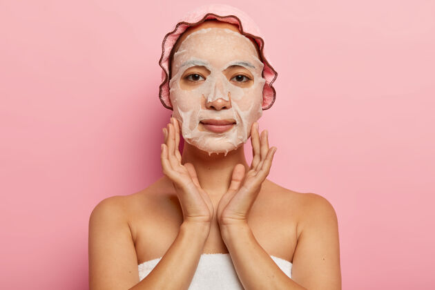 毛巾严肃的日本女性在脸上贴上滋养面膜 在皮肤上涂上保湿单品 戴上浴帽 对着粉色墙壁摆姿势女性化 美容和spa治疗理念肩膀面膜保持