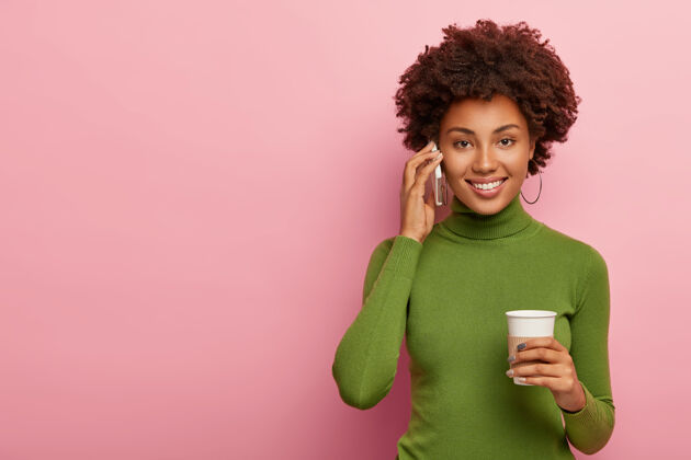 表情漂亮迷人的非洲发型女人 通过手机打电话给朋友 喝外卖咖啡 谈吐愉快 微笑愉快 谈论好消息 穿着休闲绿色套头衫 在室内摆姿势女人女性快乐