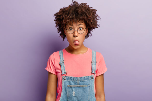 有趣有趣的黑皮肤女人的照片伸出舌头 做鬼脸 有非洲发型 戴着圆的大眼镜 牛仔工作服 粉色t恤 孤立在紫色的墙上面部表情概念非洲青少年面部