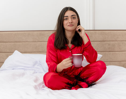相机穿着红色睡衣的年轻美女坐在床上 端着一杯咖啡 看着摄像机 在卧室的室内灯光背景下微笑着卧室坐着杯子