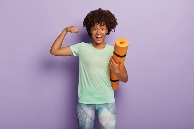 锻炼有趣的黑皮肤女人举起手臂 在训练后展示肌肉 抱着卡雷马特 和教练定期在健身房锻炼 穿着运动服 隔离在紫色的墙上力量概念卷曲积极放松