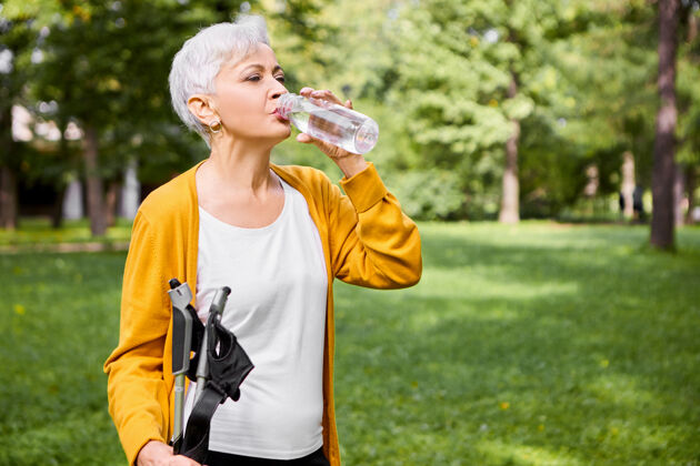 成熟夏日写真：60多岁疲惫的白发白人女性 喝着塑料瓶里的水 运动后精神焕发 拿着北欧手杖在户外摆姿势活跃瓶子老年人