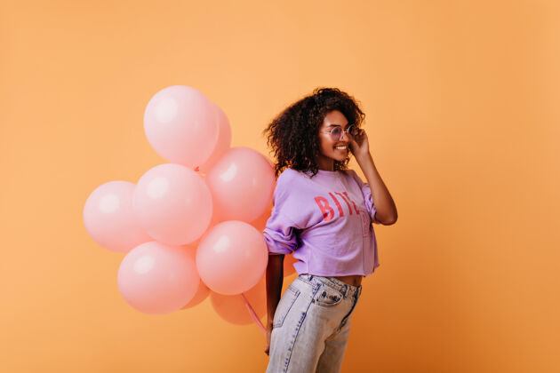 可爱戴着眼镜的时髦卷发女人手里拿着粉色的氦气球 笑着高兴的非洲生日女孩孤立在橙色上惊喜表情生日
