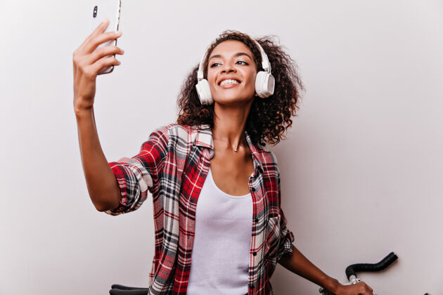 年轻灵感来自非洲白衣女士的耳机 为自己拍照穿着格子衬衫的感兴趣的女模特自拍 表情愉快自拍手机声音
