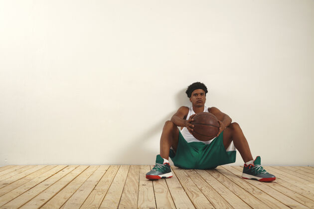 娱乐年轻严肃的篮球运动员 穿着绿色短裤和白色衬衫 手里拿着一个脏兮兮的篮球 在白墙上休息脸户外竞技