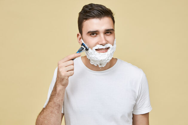 刀片一个穿着白色t恤的帅哥拿着剃须刀对着胡子刮胡子 微笑着避免皮肤发炎 注意自己的外表阳刚 时尚 美丽新鲜年轻胡茬