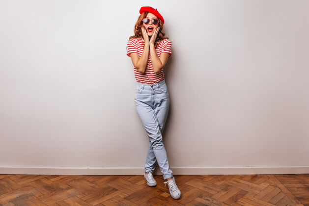 帽子穿着法国贝雷帽的幽默女孩的全身肖像穿着牛仔裤的漂亮欧洲模特在白墙上微笑Swoosh波浪发配饰